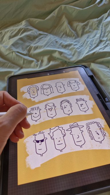 A person's hand using a stylus on a tablet to draw cartoon character faces, with musical notes indicating sound above one of the heads, resting on a green bedsheet.
