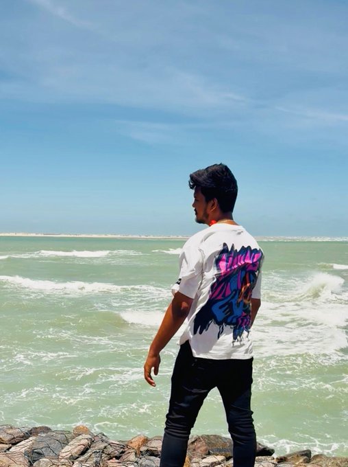 A young man in a white graphic t-shirt with pink and blue artwork standing on rocks overlooking the ocean on a sunny day.
