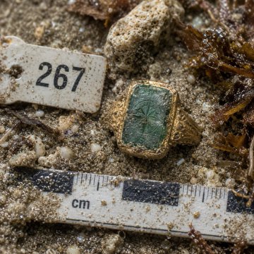 An archaeological photograph showing a gold ring with a green rectangular stone bearing a carved cross, photographed in sand next to a scale bar (marked 'cm') and a tag labeled '267'.