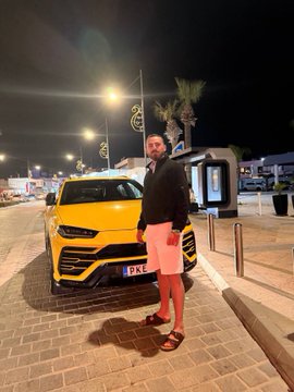 A man in shorts and sandals stands beside a brand-new yellow Lamborghini Urus (license plate "LK") at night, posing in front of it. The flashy supercar serves as a visual symbol of the post's message about rapid financial transformation.