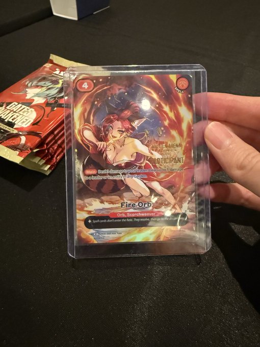 A hand holds up a holographic 'Fire Orb' trading card featuring a red-haired anime character, with 'United Fairyland' booster packs visible in the background.