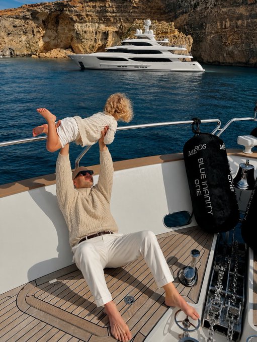 A man in a tan sweater lifts a laughing child overhead on a yacht deck, with a white superyacht and 'BLUE INFINITY ONE' fenders visible in the background.