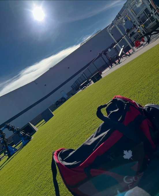 A red and black sports duffel bag with a maple leaf logo rests on green grass under a sunny sky, with a large building visible in the background.