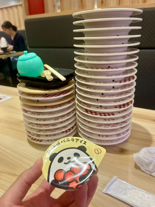 A person displays a small yellow panda-themed badge featuring "#16パニラアイス" (Panda Ice Cream) at Sushiro, with tall stacks of empty sushi plates and a green tea cup visible on the table, likely showcasing their meal progress after ordering.