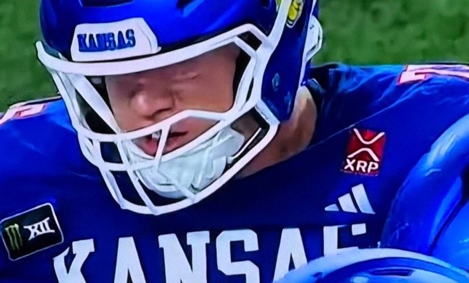 A Kansas football player, wearing jersey number 12, is shown in a close-up with a determined expression, sporting a blue helmet and jersey that prominently features the XRP logo on the sleeve. The image connects to the post by showing the XRP sponsorship on the Kansas jersey, which the author proudly highlights as a noteworthy achievement for the cryptocurrency.
