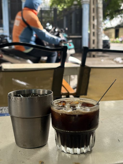 A glass of iced coffee with a black stirrer next to a metal cup of ice on a café table, with a person in an orange jacket riding a motorbike visible in the blurred background.