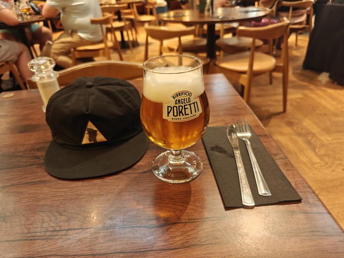 A Birrificio Angelo Poretti beer sits on a wooden table alongside cutlery and a black cap featuring a small triangular "APE" patch, suggesting the traveler is enjoying a refreshing break from the 34°C London heat at the airport.