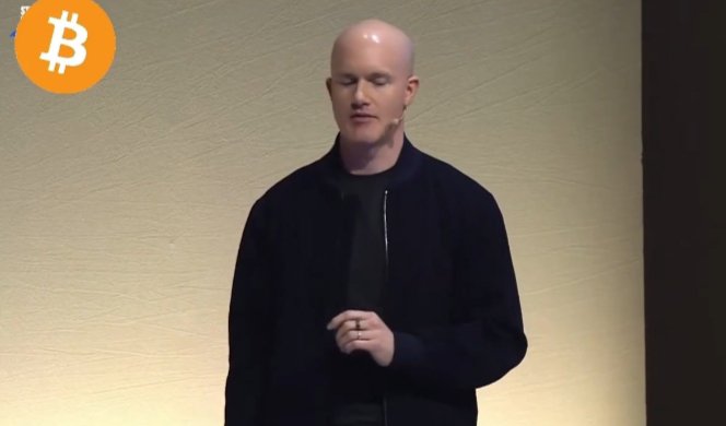 A bald man in a black jacket stands on a stage with a small Bitcoin logo in the upper left corner, appearing to give a presentation. The image accompanies an article about Coinbase CEO Brian Armstrong's belief that Bitcoin could become the world's next reserve currency amid concerns over government debt and declining confidence in traditional currencies.