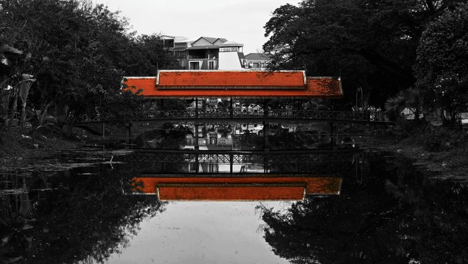 A black-and-white photograph with a selectively colored red-tiled roof of a covered bridge or pavilion reflected in calm water, surrounded by lush trees.