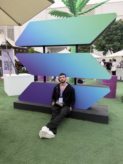 A man wearing a lanyard poses seated in front of a large installation featuring three stacked panels in a purple-to-teal gradient, with QR codes and signage for "TikTok for Education" visible nearby at an outdoor event. The colorful display and casual vibe match the upbeat "GM" (good morning) greeting.