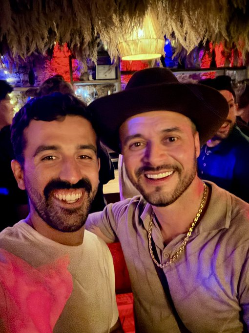 Two smiling men pose for a photo in what appears to be a bar or restaurant with a thatched roof and colorful lighting; the man on the right wears a cowboy hat and a thick gold chain.