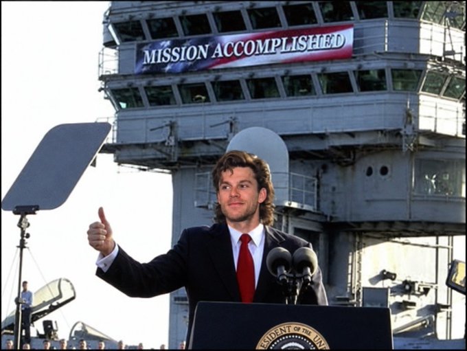 Actor Patrick Bateman (played by Christian Bale) gives a thumbs-up at a podium in front of an aircraft carrier with a 'MISSION ACCOMPLISHED' banner.