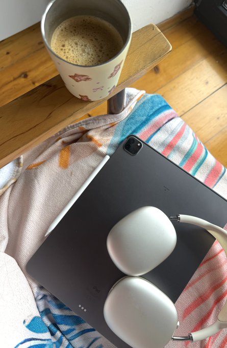 The image features an arrangement of objects that includes a laptop computer with earbuds plugged into it, a cup of coffee or tea, and a pillow. The scene takes place near a window, suggesting a cozy workspace setup. A chair can be seen in front of the window, ready for someone to sit down and work on their tasks while enjoying some background noise from the rain outside.