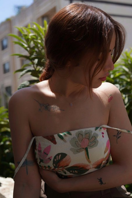 A young woman with reddish-brown hair looking downward, wearing a floral-patterned strapless top, with a small cherub tattoo visible on her chest and other tattoos on her arms, photographed outdoors in bright sunlight.