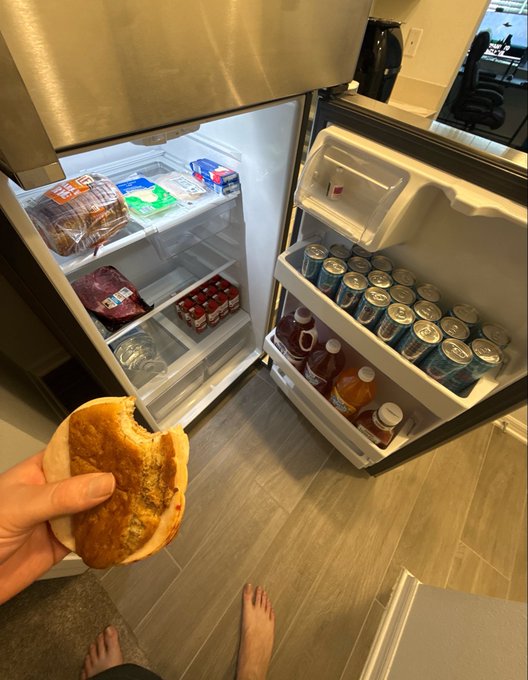 A hand holds up a half-eaten pancake in front of an open fridge stocked with various beverages in cans and bottles, along with some packaged foods and condiments on the shelves. The modest contents appear to reflect a $93 grocery haul, suggesting a casual, budget-friendly spread of mostly drinks and a few basic items.