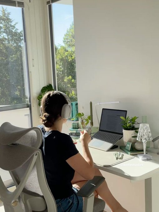 A woman with dark hair wearing headphones sits at a desk in a sunlit room with large windows, working on a laptop with code visible on the screen, surrounded by plants and a glass lamp.
