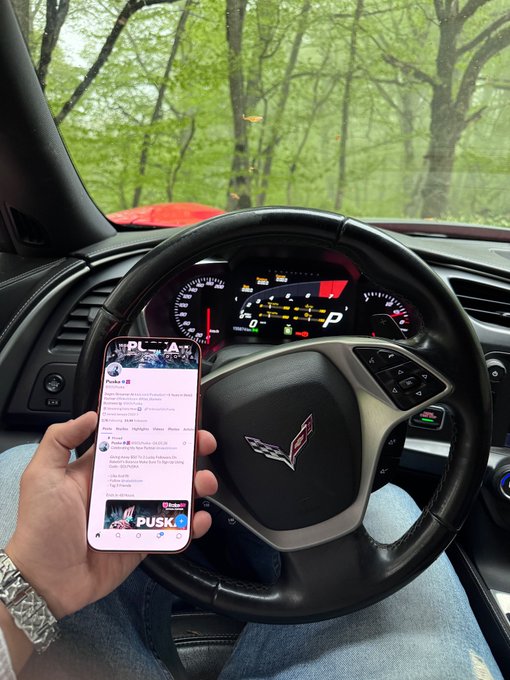 The image shows a person holding up their phone, which displays a social media app with a message that reads "Gm! Say It Back and Get $$$ Drop Your Wallets". The person appears to be sitting inside a car, possibly on a road trip or driving through an area with trees. the background of the photo is blurred, but it can be inferred that they are in a forested area.