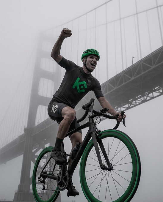 A cyclist wearing a black and green kit with a matching green helmet triumphantly raises his fist while riding a black and green bicycle, set against a foggy backdrop of a large suspension bridge, capturing the moment of ultimate victory achieved for a noble cause.