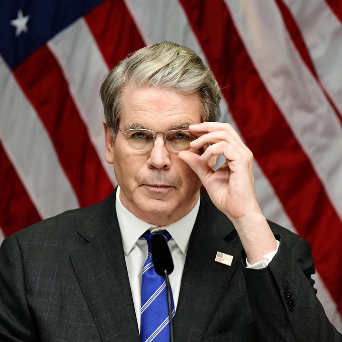 Scott Bessent, the U.S. Treasury Secretary, adjusts his glasses while speaking in front of an American flag. The image accompanies his claim that stablecoins could strengthen U.S. dollar dominance by enabling borderless, 24/7 dollar distribution without traditional banks.