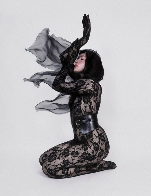 A woman kneels in a sheer black lace bodysuit with a flowing chiffon scarf, wearing a leather corset belt that emphasizes her silhouette against a stark white background. The image pairs with "gn degens" as an aesthetic, edgy fashion post showcasing avant-garde style for late-night social media engagement.