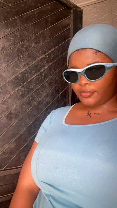 A woman wearing a light blue headscarf, matching top, and gray cat-eye sunglasses, taking a selfie with a cross necklace visible on her chest.