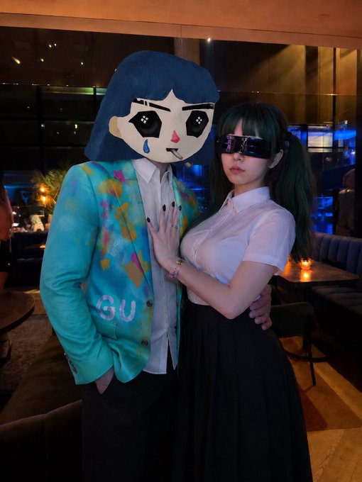 Two people pose together at an upscale lounge: one wears a turquoise Gucci (GU) graffiti-patterned blazer with an oversized cartoon mask featuring teary, star-shaped eyes, while the other sports a white blouse, black pleated skirt, and futuristic visor goggles. The lavish nightlife setting and couture fashion visually embody the "living the good life" lifestyle alluded to in the caption about investing in @ourbit.