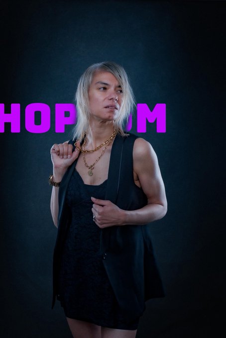 A person with blonde hair posing in a black vest and dress with a gold chain necklace, with pink text reading "HOP" and "IM" visible in the background.