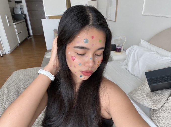 A young woman with long dark hair sits in a bedroom, her face decorated with colorful, small heart and star-shaped stickers, eyes closed in a serene expression.