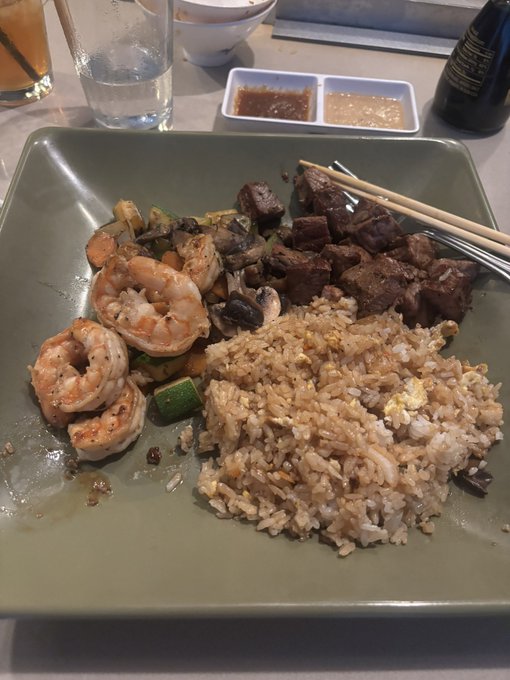 A square green plate containing hibachi-style food with shrimp, beef, mushrooms, zucchini, and fried rice, accompanied by chopsticks and two small dishes of dipping sauces.