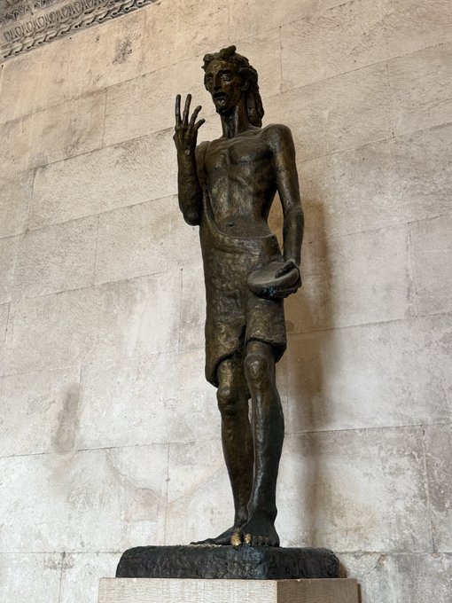 A bronze statue of a slender, bearded male figure with one hand raised, displayed on a pedestal against a stone wall.