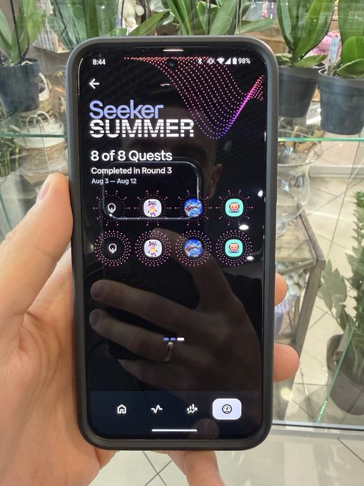 A hand holding a smartphone displaying the 'Seeker SUMMER' app screen showing '8 of 8 Quests Completed in Round 3, Aug 3 – Aug 12' with quest badges, with plants visible in the background.