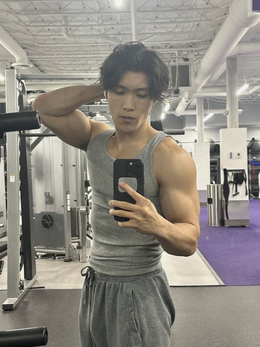 A young man in a gray tank top takes a mirror selfie at the gym, flexing his arm to show off his muscular physique. The image relates to the post's "retirement" announcement as a final pose showcasing his "chudmaxxing" physique before moving on to a new platform.