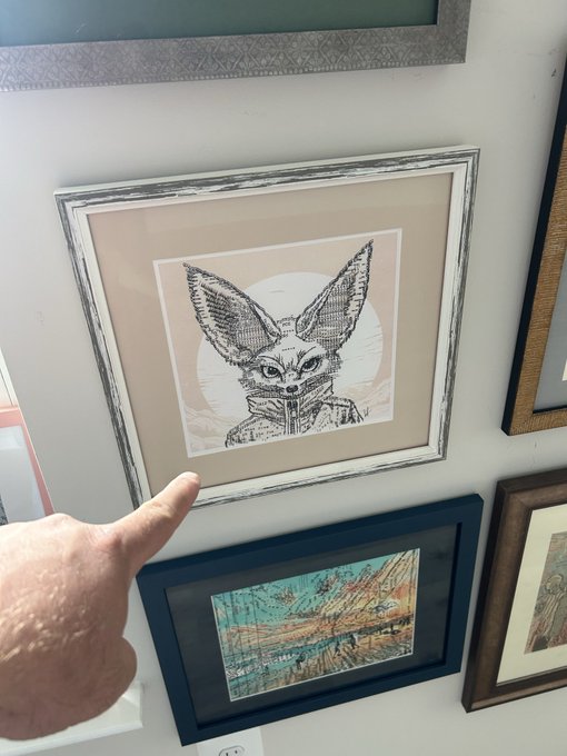 A hand points at a framed black-and-white pen drawing of a fennec fox wearing a collared jacket, displayed on a wall among other artwork including a vintage map.