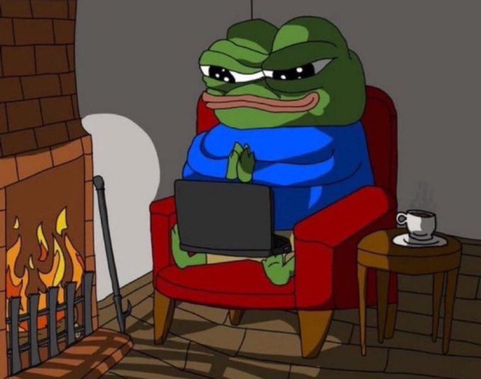 The image features a cartoon frog sitting comfortably in an armchair, using a laptop computer placed on its lap. The scene takes place near a fireplace with a cup of coffee nearby. The frog appears to be engaged in some activity or work on the laptop while enjoying the warmth and ambiance provided by the fire.