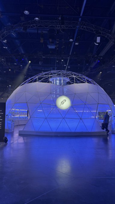 A geodesic dome lit in blue sits in a large exhibition hall, displaying a glowing circular logo with a stylized 'Q' or 'D' on its front.