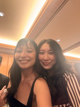 Two young women smile together for a close-up selfie in what appears to be an indoor venue.
