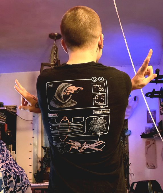 A person photographed from behind wearing a black graphic t-shirt featuring a crescent moon, brain diagrams, and text including "paranoid," "individuals," and "carnival of chaos," making peace signs with both hands.