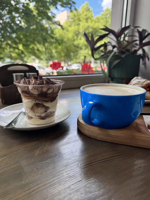 Cafe table scene with a blue ceramic coffee cup and a layered tiramisu dessert in a glass, set against a window with greenery and red flowers outside.