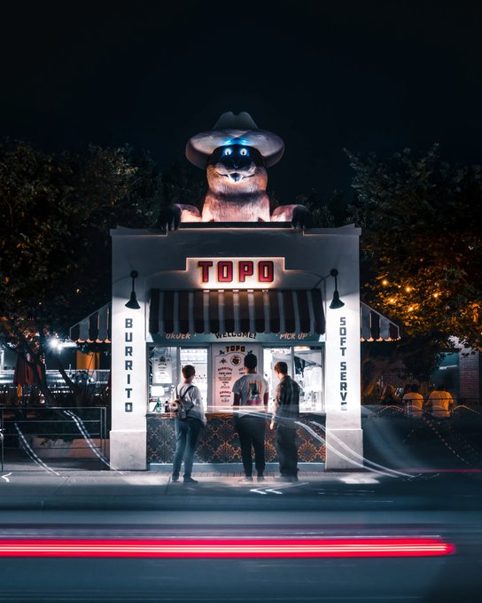 A nighttime photograph of a small 'TOPO' burrito stand with 'BURRITO' and 'SOFT SERVE' written on the sides, featuring a large mascot figure in a cowboy hat on the roof and customers gathered at the counter.