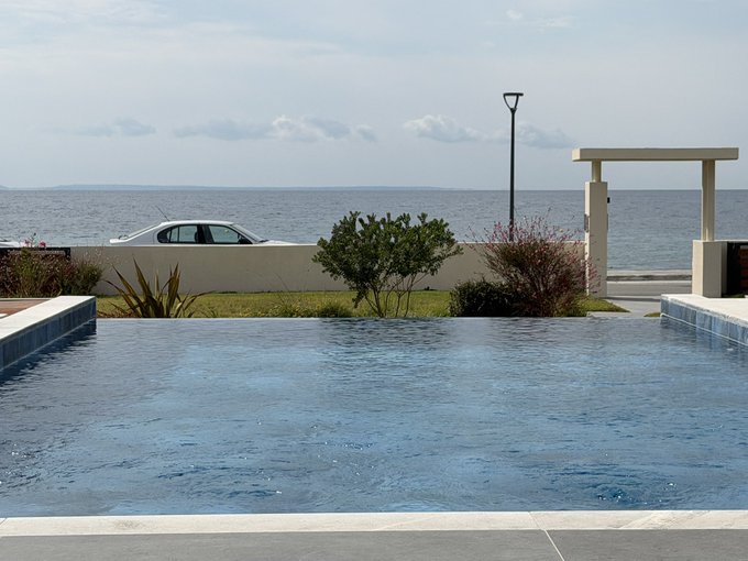 A serene infinity pool overlooks a calm body of water, with a white car parked along the road behind it and a street lamp standing against the horizon.