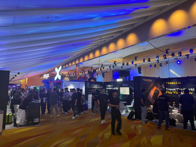 A crowded convention hall features travel-related booths with signage for 'TRAVELS.io', 'TravelPay', 'BBS', and 'Groov', illuminated by blue and orange ceiling lights.