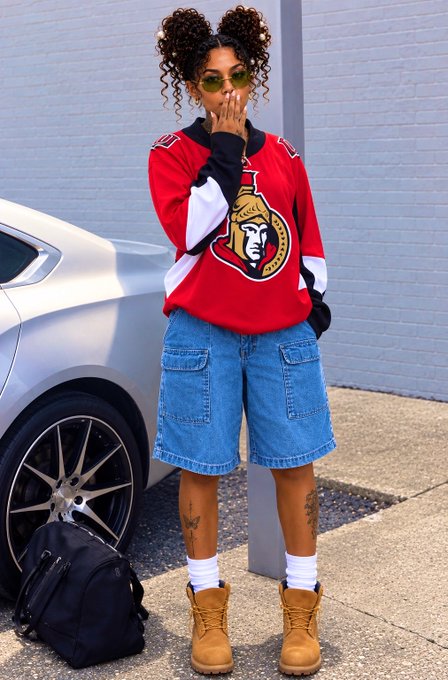 A person poses by a silver car wearing a red Ottawa Senators hockey jersey, denim shorts, tan work boots, and round sunglasses with their hair in two buns.