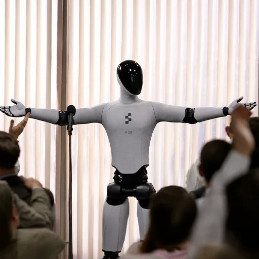 A humanoid robot with outstretched arms stands before an audience, displayed against a light-colored curtain backdrop.