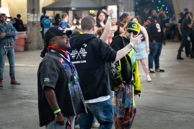 A group of people stand and dance in a busy outdoor or event setting, with one man in a cap smiling toward the camera.