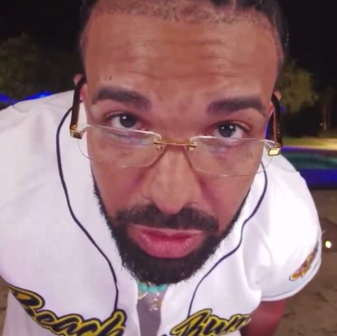 Close-up selfie-style photo of a bearded man wearing rimless glasses and a white baseball jersey with yellow trim, appearing to be outdoors at night.