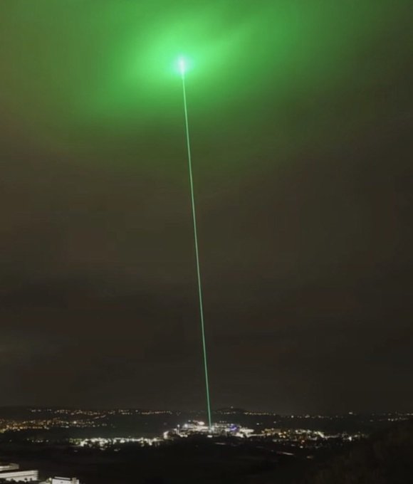 A bright green laser beam shoots up into the night sky from a ground location near city lights.