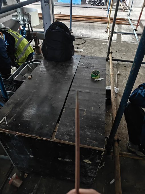 A worker holds a long metal rod in front of a worn workbench at a construction site, surrounded by scaffolding and a black backpack, capturing a moment of grueling labor. The image reflects the post's sentiment of working tirelessly through weekends while others rest, paired with crypto hashtags hinting at off-the-clock financial pursuits.