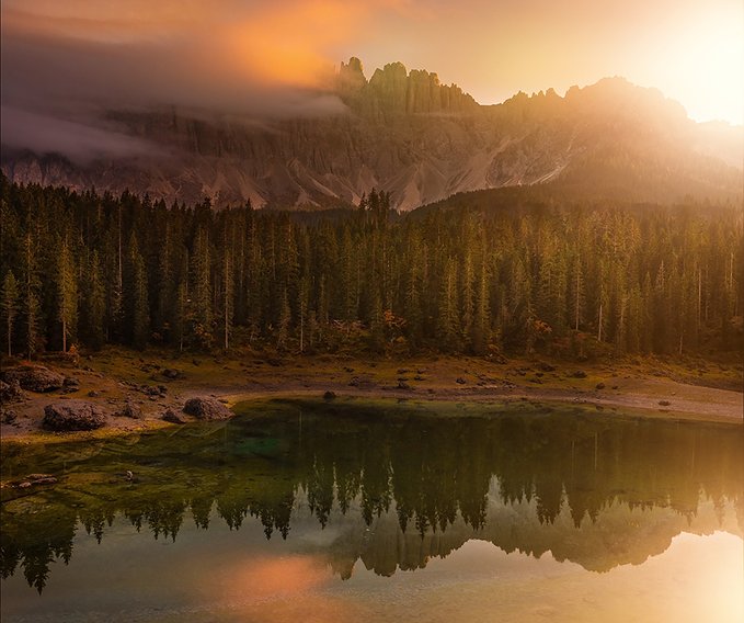 A scenic landscape featuring a calm alpine lake reflecting pine forests and jagged mountain peaks, illuminated by warm golden sunset light filtering through mist.