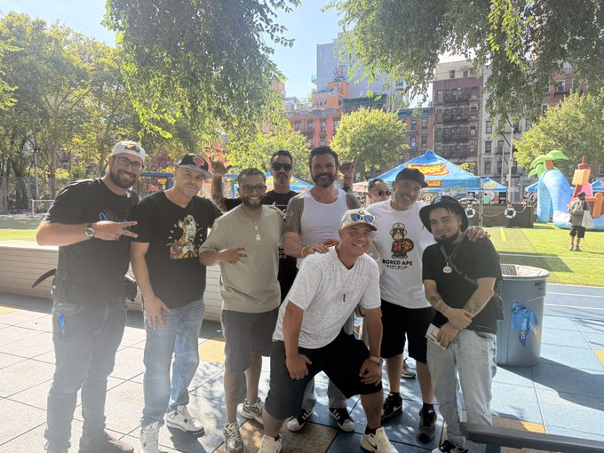 A group photo of eight men posing outdoors in what appears to be a park or plaza, with trees, buildings, and colorful tents or inflatables visible in the background on a sunny day.