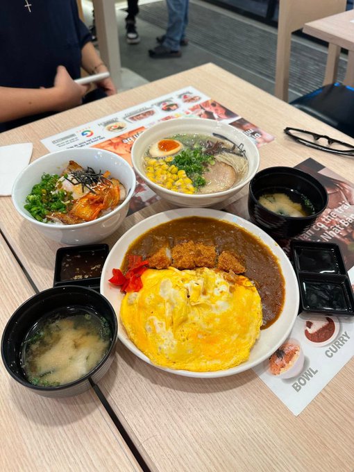 A table is set with a curry bowl topped with an omelet, a rice bowl with pork, ramen, miso soup, and side dishes at a restaurant.
